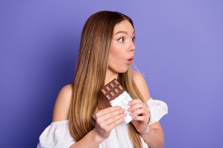 Surprised young woman holding chocolate bar against violet background, expressing amazement while enjoying sweet treatの写真素材