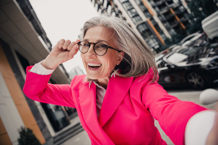 Stylish senior woman in a pink jacket smiling confidently outdoors in the citys urban streets during a sunny dayの写真素材