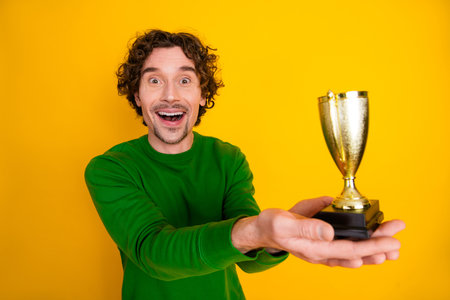 Excited young man holding a golden trophy with a vibrant yellow background, showcasing excitement and achievementの写真素材