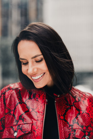 Stylish young woman in red printed jacket smiling outdoors in the city during a sunny day, vertical...