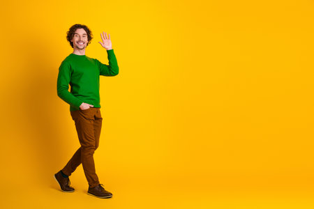 Young male with curly hair waving hand on vibrant yellow background, wearing stylish green sweater, confident and cheerfulの写真素材