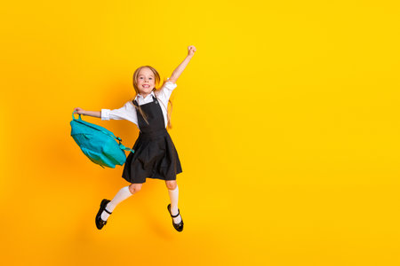 Cheerful schoolgirl in uniform jumps with joy carrying a backpack, celebrating the start of a new school year against a yellow backgroundの写真素材