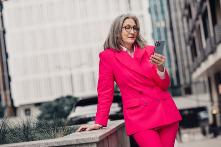 Confident mature woman in pink suit holds smartphone while standing outdoors in a city urban environment during daylightの写真素材