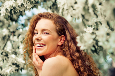 Young woman with curly hair smiling amidst blooming flowers on a sunny day, enjoying laughter and...