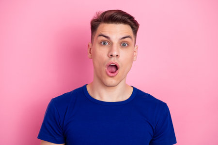 Young man with surprised expression wearing blue t-shirt standing against cheerful pink backdrop...