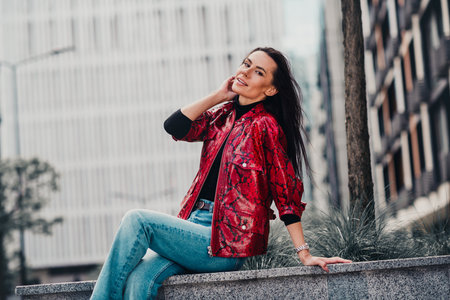 Stylish woman in red jacket enjoying a leisurely moment outdoors in an urban city setting on a sunny...