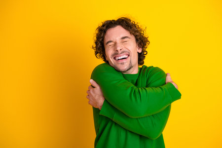 Happy young man hugging himself in green sweater on yellow background enjoying positivity and self-loveの写真素材