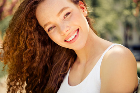 Smiling young woman with curly hair enjoying a sunny day outdoors in a charming park during...