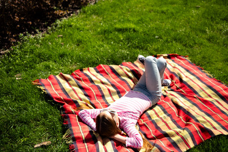 Young girl relaxing on a colorful blanket in a grassy park during a sunny dayの写真素材