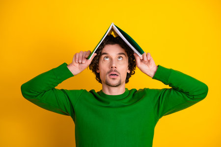 Young man holds a book on his head against yellow background wearing green sweater looking thoughtful and curiousの写真素材