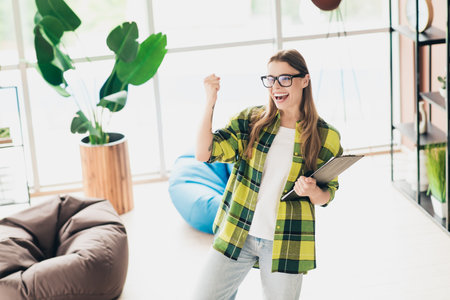 Young woman celebrating success in modern coworking space holding clipboard dressed in comfortable casual attireの写真素材