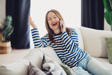 Cheerful Young Woman Talking on Mobile Phone at Home in Striped Sweatshirt Relaxing on a Comfortable Sofaの写真素材