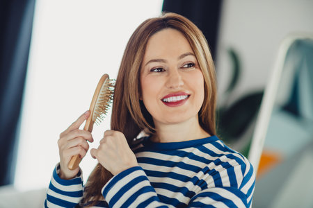Charming young woman brushing hair with a wooden brush while smiling warmly in cozy indoor setting wearing striped sweatshirtの写真素材