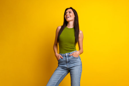 Smiling young woman in green top and blue jeans enjoying a bright yellow background, radiating positive energy and charmの写真素材