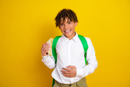 Smiling young student holding a sandwich wearing green backpack against yellow background expressing joy and school vibeの写真素材