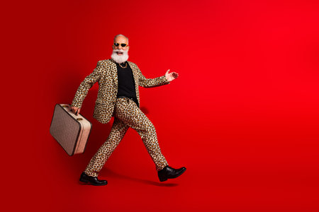 Stylish elderly man in leopard print suit holding suitcase and confidently walking on bright red backgroundの写真素材