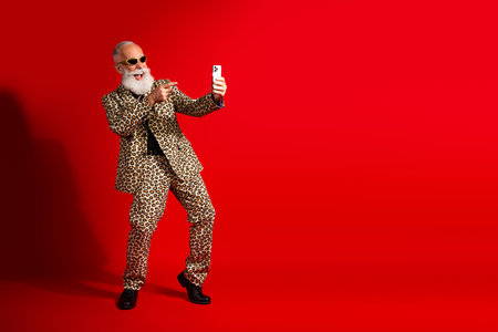 Elderly stylish man in leopard print suit posing confidently with smartphone in hand on red background, embracing charismaの写真素材