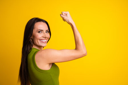 Cheerful young woman flexing her arm while smiling against a vibrant yellow background showcasing...