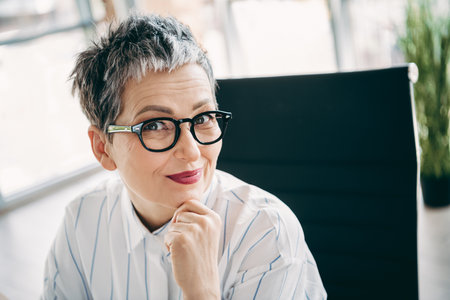 Mature businesswoman wearing glasses seated in modern office environment with professional attireの写真素材