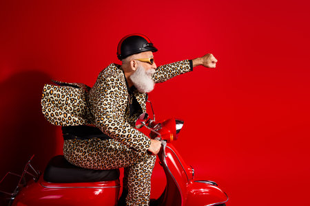 A senior man in a leopard print suit on a red scooter, exuding humor and style with a confident gesture against a bright red background.の写真素材