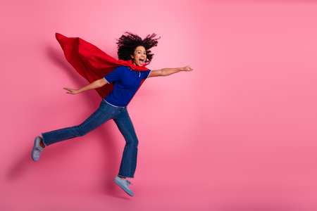 Energetic young woman in superhero pose with flowing red cape against pink background, celebrating empowerment and fun.の写真素材