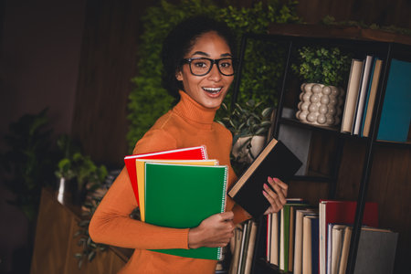 Young woman in an orange turtleneck holds books and smiles warmly while standing in a cozy library, surrounded by bookshelvesの写真素材