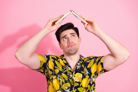 Man holding a book as a hat on a pink background wearing a tropical print shirt in a humorous poseの写真素材