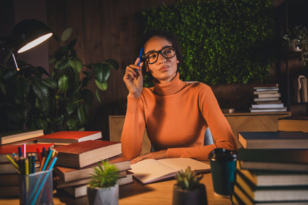Young woman in stylish turtleneck surrounded by book, pensively studying late at night in a cozy...