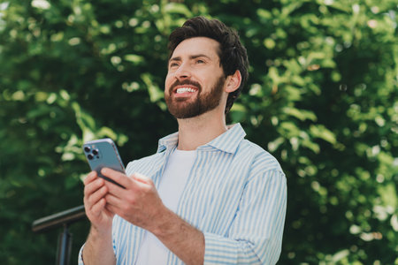 Young man outdoors smiling while using a smartphone, displaying a casual and cheerful look amidst lush greenery.の写真素材