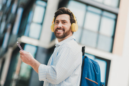 Handsome man with yellow headphones enjoying music and a phone outdoors in the city, reflecting a...