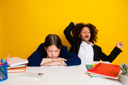 Two multicultural schoolgirls in a classroom setting, one yawning and the other resting her head,...