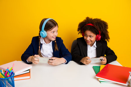 Two cheerful schoolgirls with headphones enjoying mobile games at a desk amidst colorful stationery...