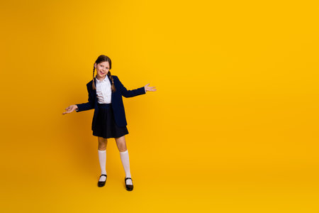 Delighted schoolgirl in uniform posing confidently with cheerful energy against a bright yellow backgroundの写真素材