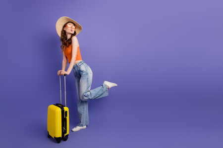 Pretty young woman posing with yellow suitcase against purple background, dressed in trendy casual outfit, ready for travelの写真素材
