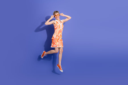 Happy woman in orange dress and sunglasses jumping against vibrant purple background expressing joy