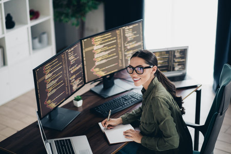 Charming female programmer working on multiple monitors at home office while writing code and taking...