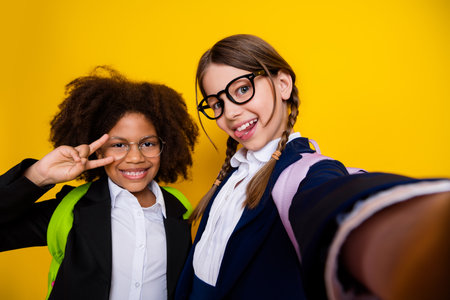 Two cheerful schoolgirls taking a selfie against a yellow background, expressing friendship and school spirit with smiles and peace signsの写真素材