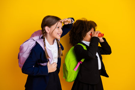 Two young schoolgirls explore with binoculars and smiles on a bright yellow background, symbolizing...