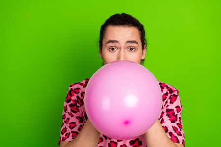 Young man blowing a pink balloon against a vibrant green background, dressed in stylish...