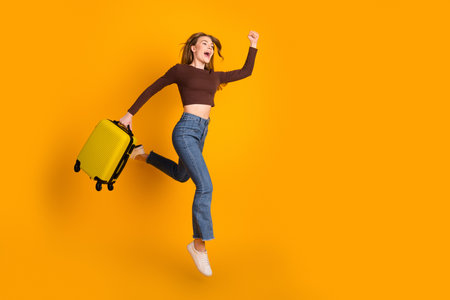 Young woman jumping against yellow background holding a suitcase, demonstrating joy and enthusiasm for travelの写真素材