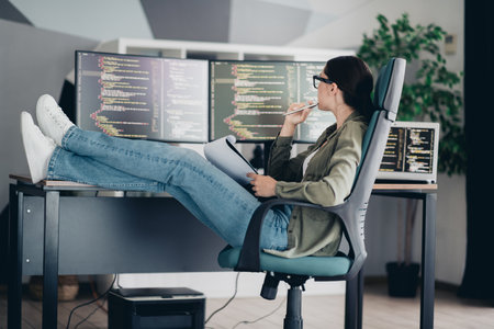 Young programmer working remotely at her computer station, analyzing code with a relaxed and focused pose in an officeの写真素材