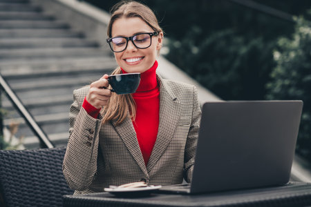 Confident businesswoman enjoying coffee in an outdoor urban location while working with her laptop on a sunny dayの写真素材