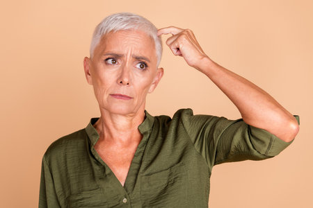 Mature woman with short gray hair gesturing thoughtfully in casual khaki shirt against beige background showcasing lifestyle and fashionの写真素材