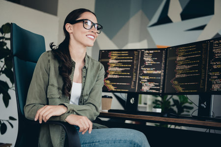 Young female programmer working at her desk in a modern workspace with dual monitors, coding and smiling confidentlyの写真素材