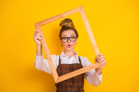 Portrait of creative student with glasses and wooden frame excited on yellow background celebrating...