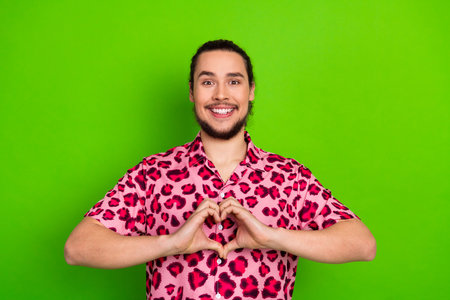 Young man in pink leopard patterned shirt making heart gesture on vibrant green background