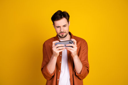 Handsome man in casual attire engrossed with a smartphone against a vibrant yellow background expressing style and modern lifestyleの写真素材