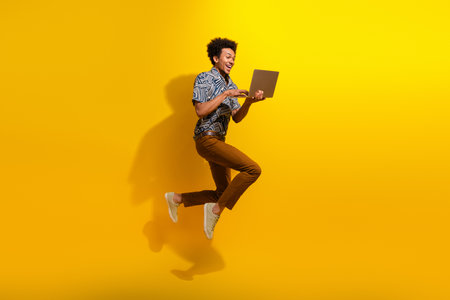 Young man jumping with a laptop against a yellow background, expressing excitement and joy in casual fashionの写真素材