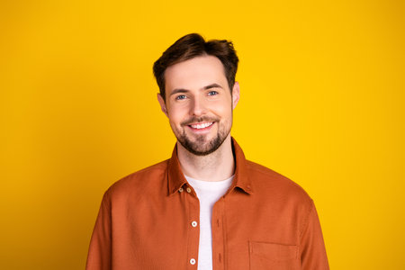 Young man wearing a casual orange shirt smiles confidently on a vibrant yellow background, exuding charm and positivityの写真素材