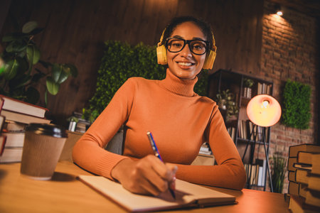 Young woman in a cozy apartment studying late at night, wearing headphones and a turtleneck, seated...